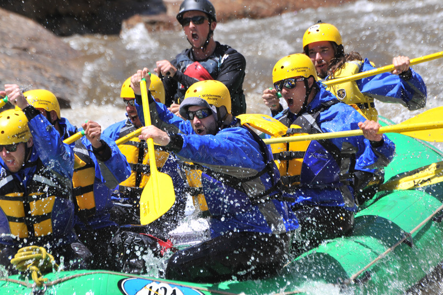 People in a raft yell in excitement as they paddle through rapids in yellow helmets