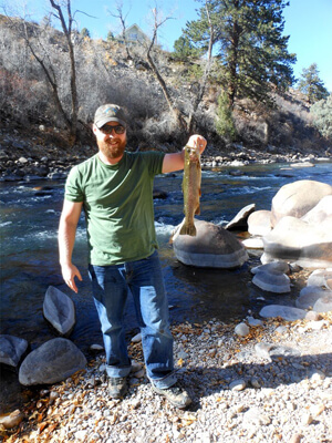 A man holds up a trout on the side of a river