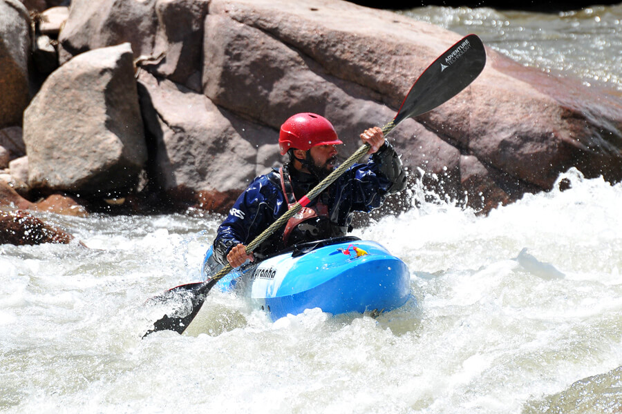 A man paddles a blue kayak in a river wearing a red helmet