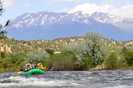 A scenic photo of a raft going down a river with a mountain range in the background