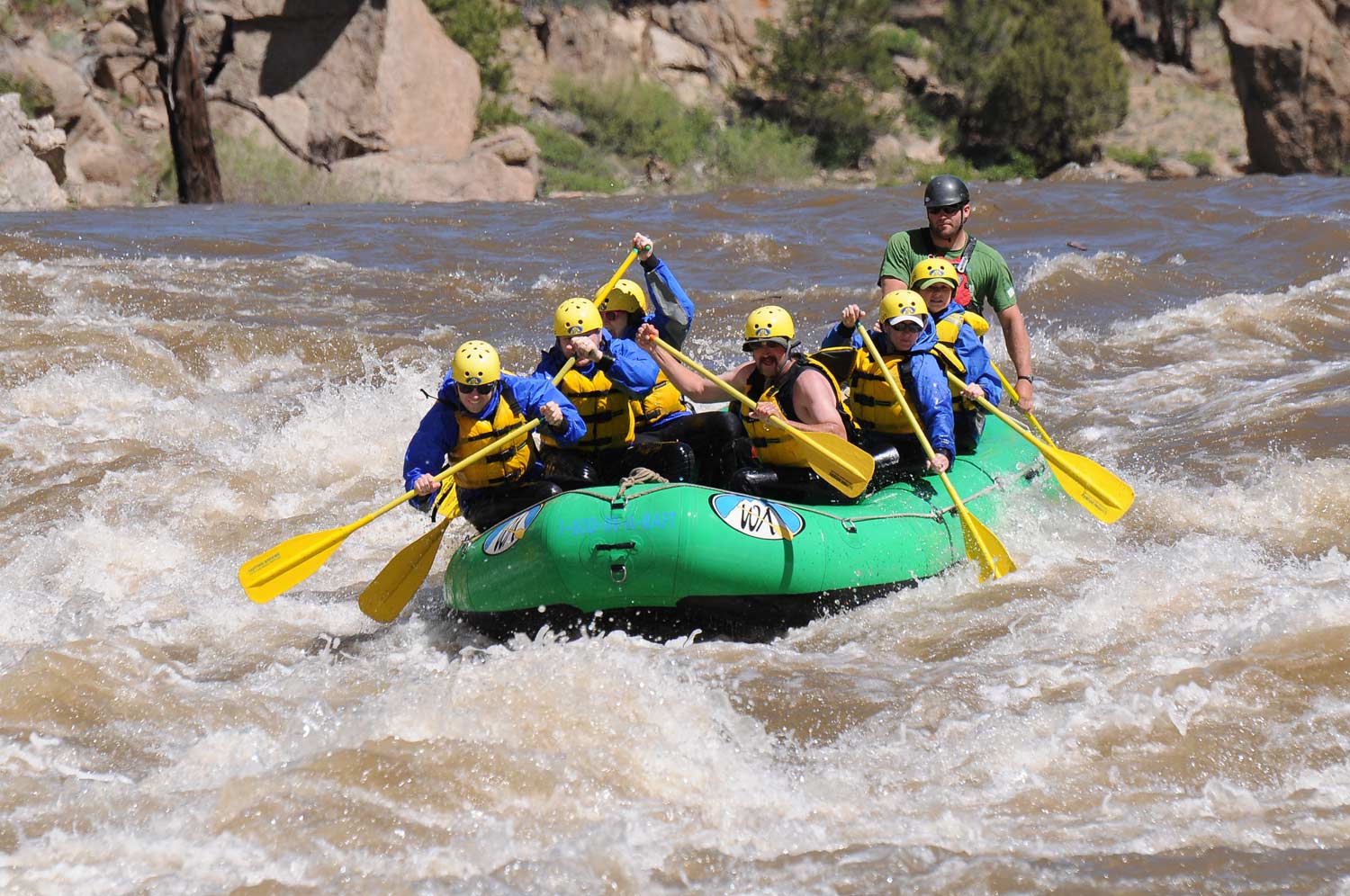 Seven people on a green raft holding yellow oars paddle down a river