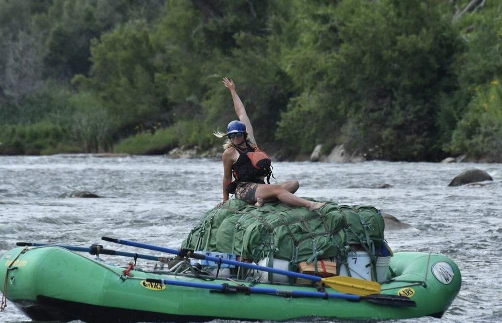 A woman site on top a bunch of gear on a green raft and poses