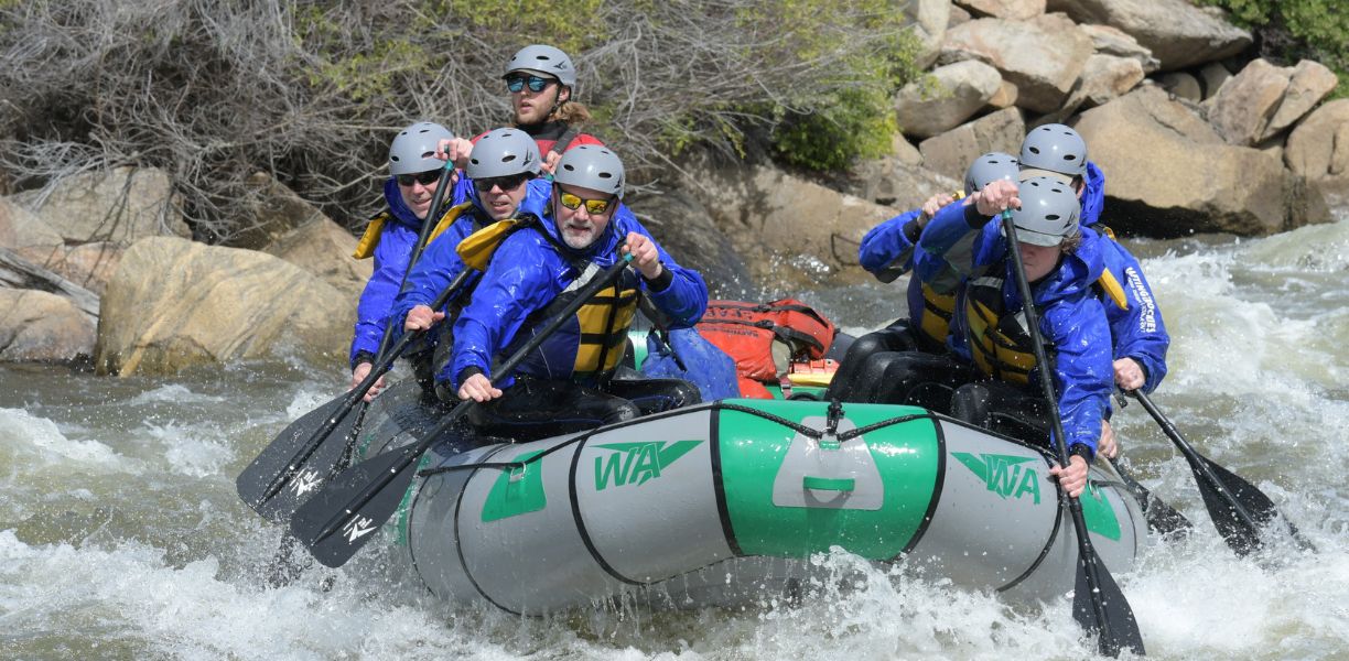 People paddle hard in a grey and green raft on the river