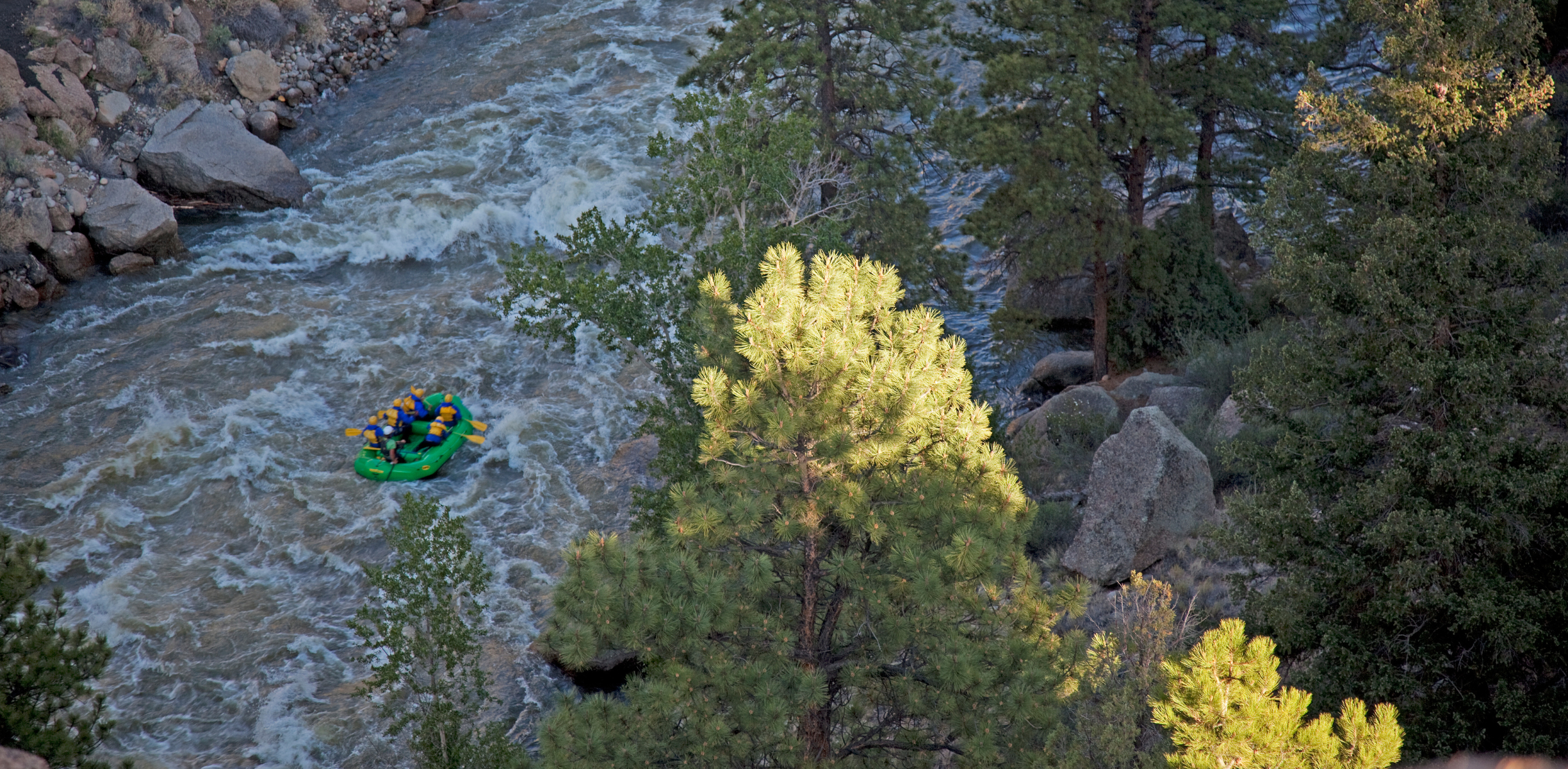 A group of people whitewater rafting in a green raft on a rocky river surrounded by trees on the Arkansa River.