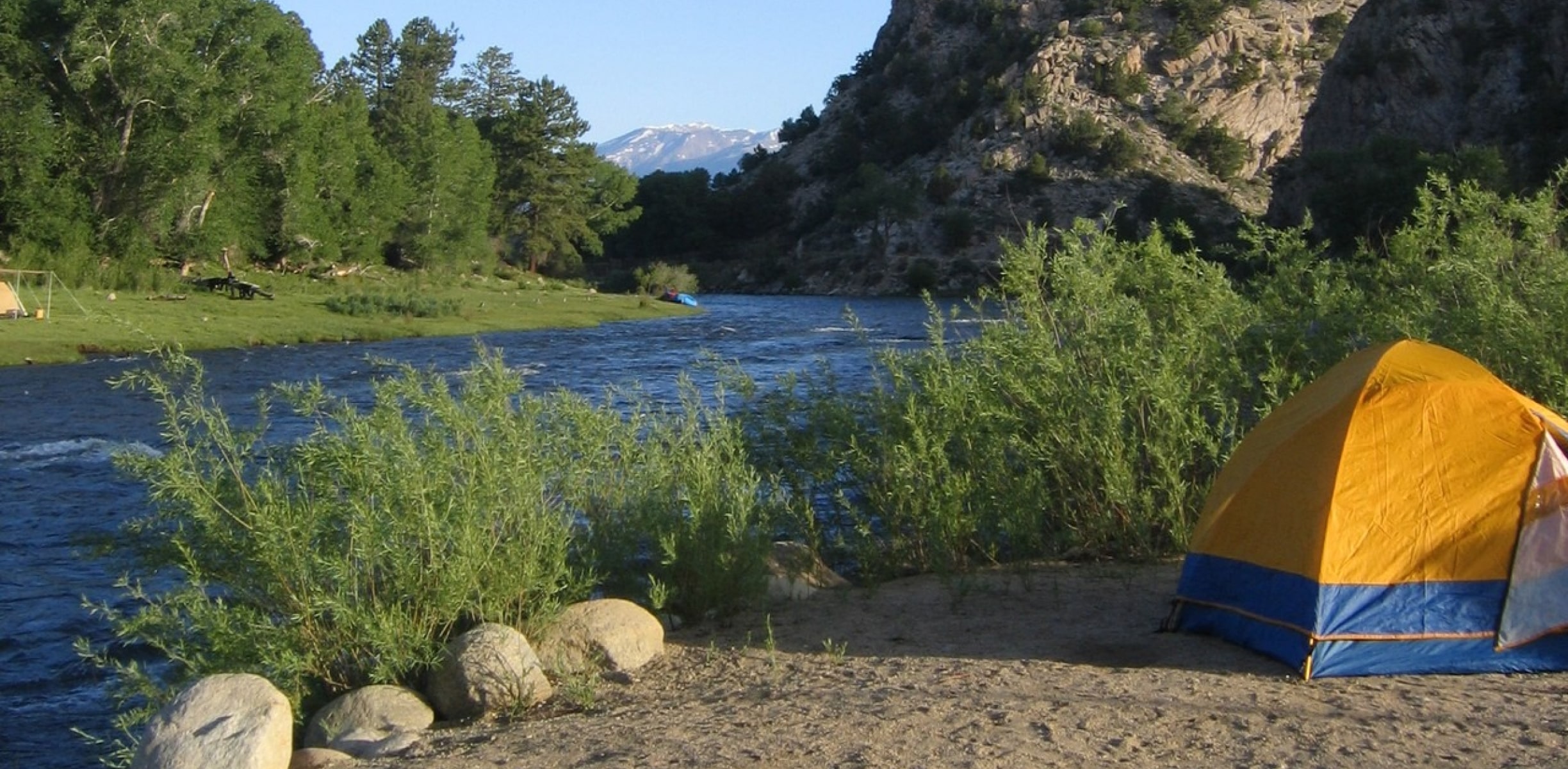 Yellow and blue tent set up beside a river with trees, rocky cliffs, and mountains in the background.